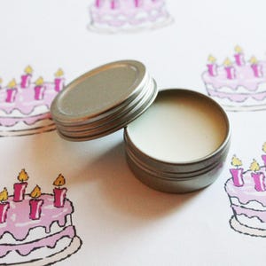 May include: An open, small, silver-colored tin of cream-colored balm next to its lid. The background features a repeating pattern of pink birthday cakes with lit candles, creating a festive theme.