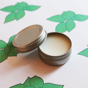 May include: A small, open, silver-colored tin of balm with a white, creamy texture. The lid is slightly ajar, next to the tin. The background features a white surface with green leaf illustrations, creating a natural, botanical theme.