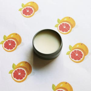 May include: A small, open, silver-colored tin filled with a light-colored substance, centered on a white surface. The surface is decorated with illustrations of halved grapefruits, each with a red interior and yellow rind.