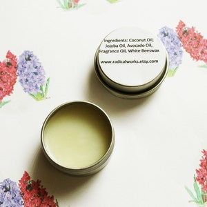 May include: A small, open, silver-colored tin containing a light green balm. The lid of the tin displays the ingredients: Coconut Oil, Jojoba Oil, Avocado Oil, Fragrance Oil, and White Beeswax. The website www.radicalworks.etsy.com is also printed on the lid. The background features watercolor floral art.