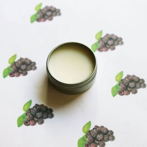 May include: A small, round, silver-colored tin filled with a light-colored balm. The tin is centered on a white surface decorated with watercolor illustrations of blackberries and green leaves.