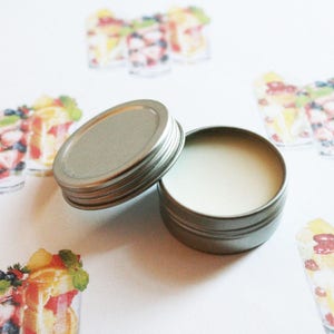 May include: An open, small, silver-colored tin with a light-colored balm inside, next to its lid. The background features watercolor illustrations of fruit parfaits in clear glasses, with strawberries, blueberries, and other fruits.
