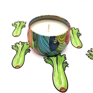 May include: A colorful candle in a tin container with a whimsical design. The candle is surrounded by five green celery stalks.