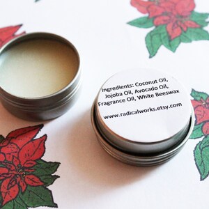 May include: Two small, open, silver-colored tins filled with a creamy balm. One tin's lid is visible, displaying the ingredients: Coconut Oil, Jojoba Oil, Avocado Oil, Fragrance Oil, and White Beeswax. The background features poinsettia illustrations.