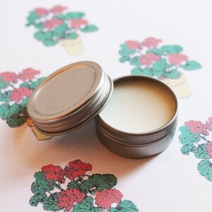 May include: An open, small, round, silver-colored tin with a screw-on lid. The tin contains a light-colored balm. The background features a repeating pattern of red and green geraniums in yellow pots.