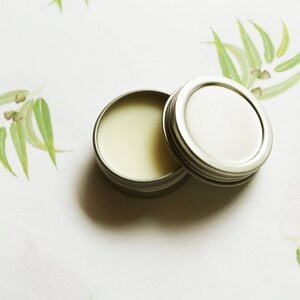May include: Open, small, round, silver-colored tin with a light yellow balm inside. The lid is next to the tin. The background is white with green and yellow leaf and branch designs.