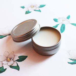 May include: A small, open, silver-colored tin with a screw-on lid, filled with a white, creamy substance. The tin sits on a white surface decorated with watercolor-style magnolia flowers and green leaves.