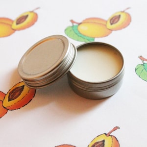 May include: An open, small, round, silver-colored tin container filled with a white substance, next to its lid. The background features a repeating pattern of illustrated apricots with green leaves.
