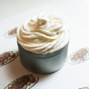 Puede incluir: Un pequeño recipiente metálico gris lleno de un remolino de crema blanca. La crema es espesa y texturizada, con picos visibles. El recipiente se encuentra sobre una superficie blanca decorada con ilustraciones de comida.