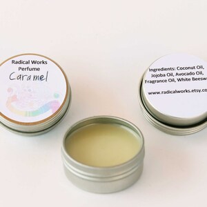 May include: Three small, round, silver-colored tins of perfume. One tin is open, revealing a light yellow balm. Two tins have white labels with text, including "Caramel" and ingredient information. The brand name "Radical Works" is also visible.