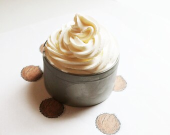 May include: A small, gray container filled with a swirl of creamy, off-white substance, possibly a cosmetic product or food item. The container is surrounded by several sketched brown circles, resembling coconuts, on a white background.