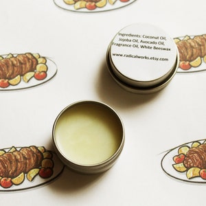 May include: A small silver tin with a white label that reads "Ingredients: Coconut Oil, Jojoba Oil, Avocado Oil, Fragrance Oil, White Beeswax www.radicalworks.etsy.com". The tin is sitting on a white surface with several drawings of a roasted meat dish with potatoes and tomatoes.