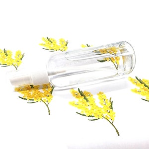 Puede incluir: Un frasco rociador de vidrio transparente con una boquilla blanca, sobre una superficie blanca. El frasco está rodeado de ilustraciones de flores de mimosa amarillas. El frasco está lleno de un líquido transparente.