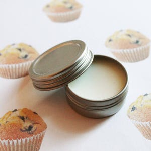 May include: An open, silver-colored tin with a lid, filled with a white substance, possibly a balm or cream. The tin is surrounded by images of muffins with blueberries. The muffins are in white paper cups.