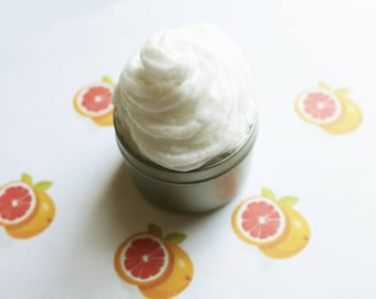 May include: A small, silver container filled with a large swirl of white, fluffy cream. The container sits on a white surface decorated with illustrations of grapefruit halves. The cream appears to be a cosmetic product.