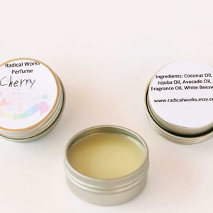 May include: Three small, silver-colored tins of perfume. One tin is open, revealing a light yellow balm. Two tins have white labels with text: "Radical Works Perfume Cherry" and "Ingredients: Coconut Oil, Jojoba Oil, Avocado Oil, Fragrance Oil, White Beeswax".