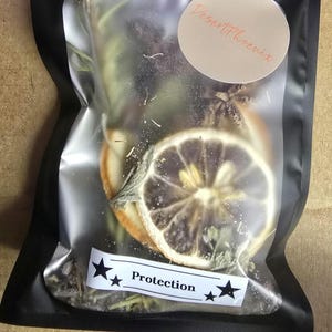 May include: A clear plastic bag filled with dried herbs, a dried orange slice, and a dried lemon slice. The bag has a white label with the word "Protection" and star graphics. A circular sticker with the text "DesertPhoenix" is also visible.