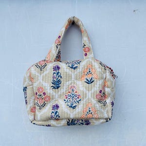 May include: A beige quilted tote bag with a floral pattern in shades of pink, orange, purple, and blue. The bag has a rectangular shape with a zipper closure and two handles.