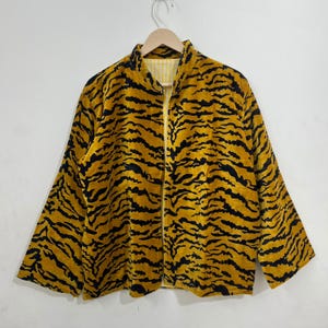 Pode incluir: Um casaco de manga comprida com um padrão de riscas de tigre em preto e amarelo mostarda. A peça tem uma gola subida, um design frontal aberto e um forro às riscas finas em amarelo claro e branco visível no interior da gola.