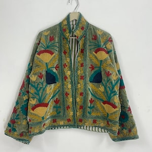 May include: A green velvet jacket with floral embroidery in yellow, red, and teal. The jacket features a high collar and a striped lining. The embroidery is detailed and covers the front and sleeves.
