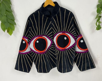 Könnte beinhalten: Schwarze Samtjacke mit hohem Kragen und langen Ärmeln. Die Jacke hat ein sich wiederholendes Augendesign mit roten, weißen und lila Akzenten und goldfarbenen Strahlen, die von den Augen ausgehen. Die Jacke hängt an einem Holzbügel.