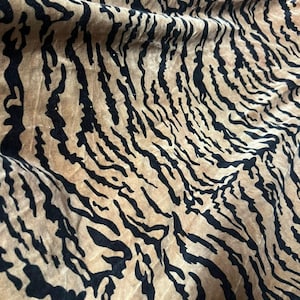 Puede incluir: Una tela con un patrón repetitivo de rayas de tigre en negro sobre un fondo beige. El material tiene una superficie suave y texturizada con pliegues y drapeados visibles que crean profundidad y sombras sobre el tejido.