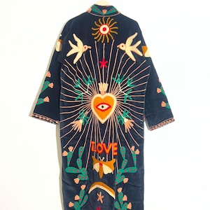 Könnte beinhalten: Ein langer, dunkelblauer Morgenmantel mit farbenfrohen Stickereien, die ein zentrales Herz mit Auge, Sonnenstrahlen, Vögel, Kakteen, einen Nachtfalter und das Wort LOVE zeigen. Das Design enthält orange, gelbe, grüne und rote Akzente auf dem Stoff.