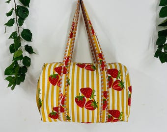 Puede incluir: Un bolso tote rectangular con un patrón de rayas amarillas y blancas, adornado con ilustraciones de fresas rojas. El bolso tiene asas a juego a rayas y es ideal para llevar objetos.