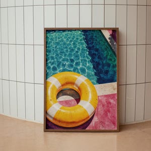 Puede incluir: Impresión enmarcada que muestra una piscina con un flotador inflable a rayas amarillas y blancas. El agua es azul oscuro y el marco es marrón. La impresión está apoyada contra una pared de azulejos blancos.