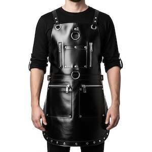 May include: Black leather apron with silver zips and metal ring details. The apron features two large front pockets and adjustable straps. The design has a modern, industrial aesthetic.