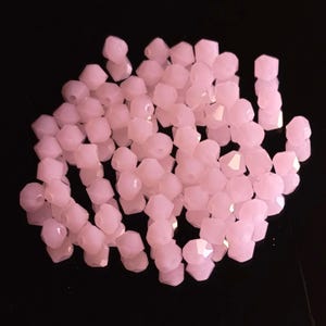 Può includere: Una collezione di perline rosa chiaro sfaccettate. Le perline hanno una forma geometrica con più lati, creando un effetto scintillante. Le perline sono sparse su uno sfondo nero, evidenziando il loro colore e la loro forma.