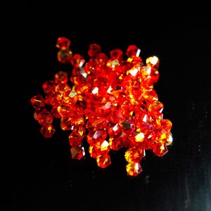 Può includere: Un mucchio di perline di cristallo rosso-arancio sfaccettate. Le perline sono sparse su una superficie nera, catturando la luce e creando un effetto scintillante. Queste perline sono adatte per la creazione di gioielli e progetti artigianali.