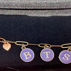 May include: A gold-colored chain bracelet with charms. The bracelet features the number "7", purple flower charms, heart charms, and purple circular charms with the letters "BTS" in white. The bracelet is on a dark surface.