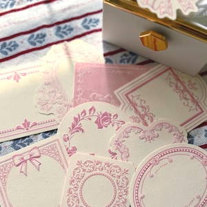 May include: A selection of pink and cream decorative cards featuring floral and bow motifs. The cards are in diverse shapes, including squares, circles, and rectangles. A white box with gold trim and a bow sticker are visible in the background.