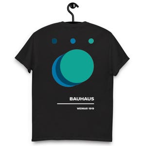 Puede incluir: Camiseta negra con un diseño geométrico turquesa y azul. El diseño incluye tres círculos pequeños encima de un círculo más grande. La palabra "BAUHAUS" y "WEIMAR 1919" están impresas debajo.