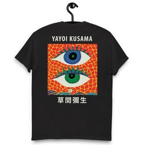 Puede incluir: Camiseta negra con un diseño gráfico colorido. El diseño incluye dos ojos estilizados con iris azules y verdes, sobre un fondo rojo y naranja. También se ve el texto "YAYOI KUSAMA" y caracteres japoneses.