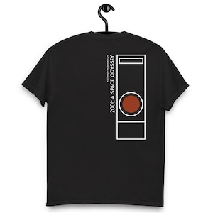 Pode incluir: T-shirt preto com um design minimalista do filme "2001: Uma Odisseia no Espaço". O design apresenta um contorno retangular branco com um círculo vermelho e o título do filme em texto branco. A t-shirt está pendurada num cabide preto.