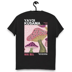 Puede incluir: Camiseta negra con un diseño de setas coloridas en rosa, rojo, amarillo y verde, con el texto "YAYOI KUSAMA TOKYO 1998" y texto japonés. El diseño incluye setas sobre un fondo geométrico rosa. La camiseta está colgada de una percha negra.