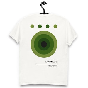 Puede incluir: Camiseta blanca con un diseño de círculos concéntricos verdes y cuatro puntos verdes encima. La palabra "BAUHAUS" está impresa debajo, con "17 JUNE 1921" debajo. La camiseta está colgada en una percha negra.