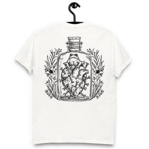 Puede incluir: Camiseta blanca con una ilustración en blanco y negro. El diseño muestra una rana dentro de un tarro de cristal, sentada sobre un corazón atravesado por clavos. Elementos florales rodean el tarro, creando un diseño artístico único.
