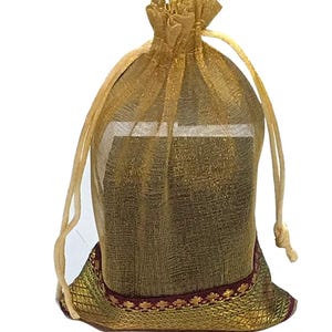 May include: A gold-colored, sheer drawstring bag. The bag is filled with an unseen object. The bottom of the bag has a decorative band with a red and gold pattern. The drawstring is a light beige color.