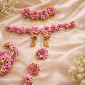May include: A set of floral jewellery, including a headpiece, earrings, and a ring, displayed on a satin surface. The pieces feature pink flowers with yellow accents and dangling pearl chains. The jewellery is designed for special occasions.