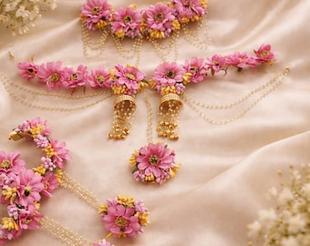 May include: A set of floral jewellery, including a headpiece, earrings, and a ring, displayed on a satin surface. The pieces feature pink flowers with yellow accents and dangling pearl chains. The jewellery is designed for special occasions.