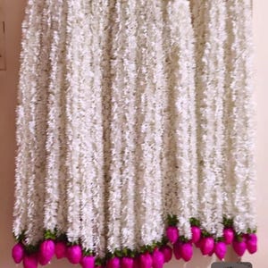 May include: White floral garlands with a textured appearance, hanging vertically. The garlands have a green base with vibrant pink, teardrop-shaped accents at the bottom. The overall aesthetic is delicate and decorative, suitable for events or home decor.