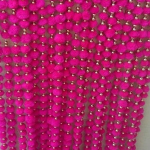 Könnte beinhalten: Eine leuchtend pinkfarbene Girlande aus flauschigen Pompons und kleinen goldenen Perlen. Die Girlande hängt vertikal, mit dekorativen goldfarbenen Glocken am unteren Ende. Diese festliche Dekoration eignet sich für Feiern.