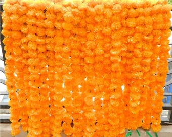 May include: A dense curtain of vibrant orange marigold flowers arranged in multiple vertical strands. Each strand consists of numerous ruffled, round blossoms strung together to create a full, textured, and uniform floral display.