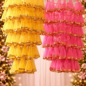 May include: Two decorative hanging lamps, one yellow and one pink, are suspended from the ceiling. Each lamp features multiple tiers of cone-shaped fabric with gold trim. The background is softly lit with bokeh lights.