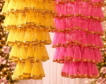 May include: Two decorative hanging lamps, one yellow and one pink, are suspended from the ceiling. Each lamp features multiple tiers of cone-shaped fabric with gold trim. The background is softly lit with bokeh lights.
