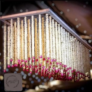 Puede incluir: Un marco rectangular de madera adornado con largas hebras de flores blancas, acentuadas con vibrantes flores rosas en la parte inferior. El arreglo floral cuelga del techo, creando una exhibición decorativa.