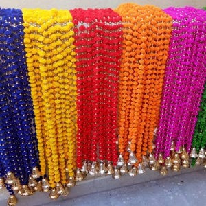 Puede incluir: Una colección de guirnaldas coloridas en tonos azul, amarillo, rojo, naranja, rosa y verde. Cada guirnalda está hecha de pequeños elementos redondos y texturizados y adornada con borlas doradas en forma de campana en la parte inferior. Las guirnaldas están dispuestas una al lado de la otra.